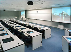 Classroom with fixed desks featuring under-top monitors and presentation screens and board at front of room
