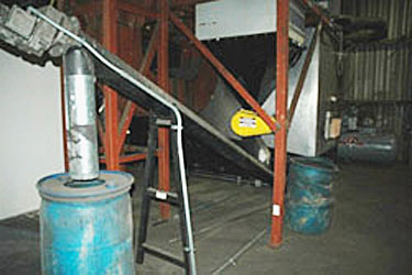 Photo of an automated ash-removal system on a wood-fire boiler. The boiler is located in the lower right portion of the photo, and the system uses an auger to remove ash from the boiler. It then delivers the ash to a blue barrel located on the left of the photo.