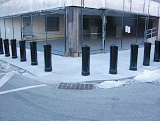 bollards on along a sidewalk and around a corner with a grate on the road in front and some snow on the ground and a new construction building behind