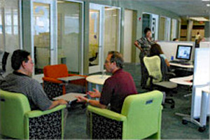 two men in upholstered chairs having a meeting alongside workers at thier workstations/cubicles
