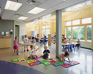 Young children playing on floor mats and at tables in a well-lit, window-filled child development center