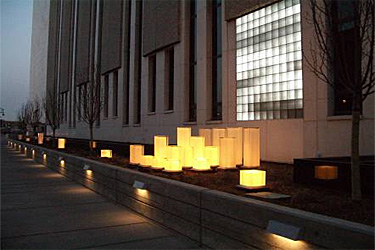 Sidewalk view of building at night with lights embedded in short wall illuminating the sidewalk and array of decorative lights in front of glass block facade