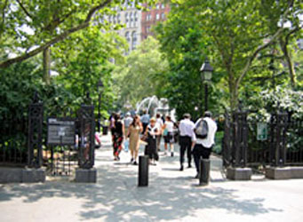 An open entry gate with pedestrians walking along a path between trees set in low walls and among bollards