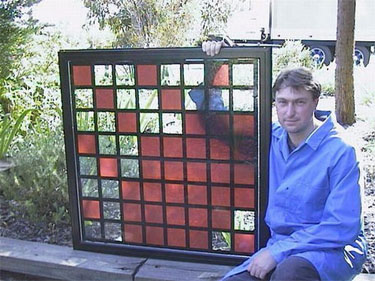 Photo of a man holding a window panel that contains a grid comparing cells that are colored with solar dye against squares with clear glass